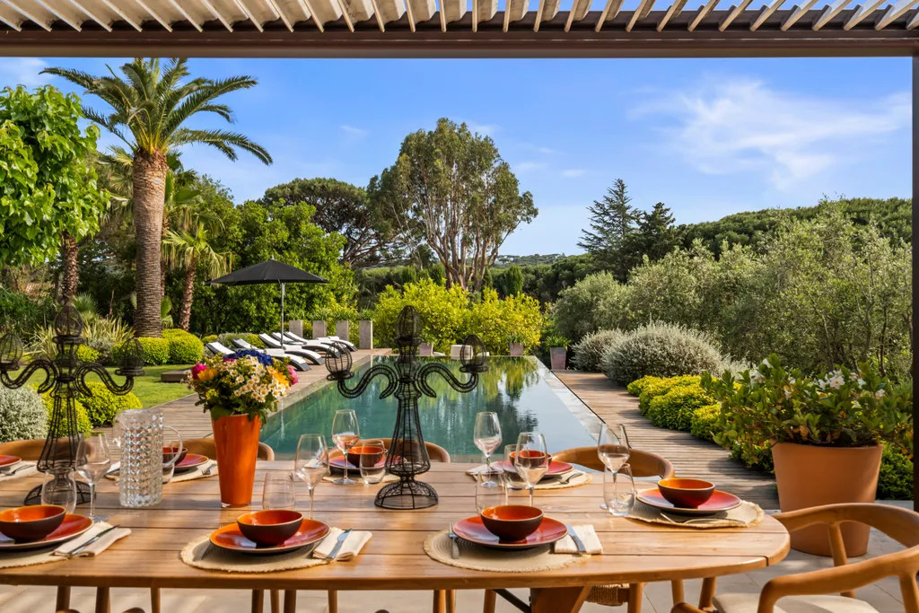 An outdoor table dressed with terracotta plates and flowers, set under the pergola above the pool