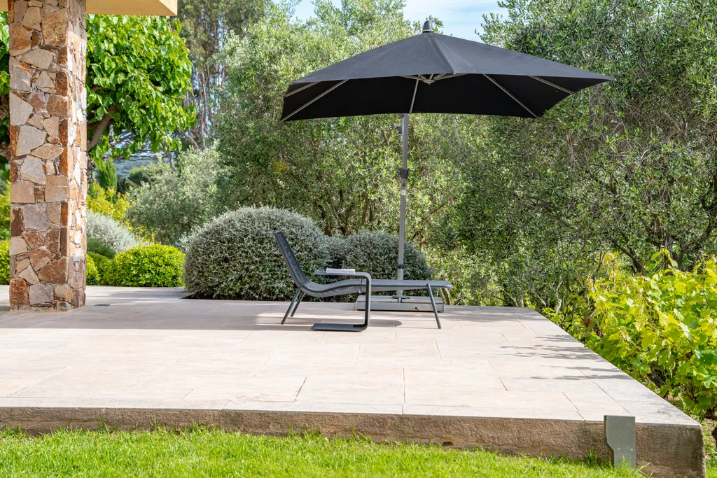 A shaded stone terrace with a lounge chair beneath a parasol, edged by clipped greenery