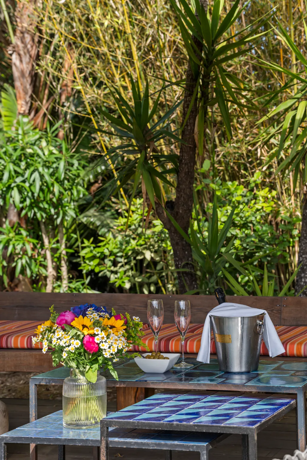 Chilled rosé and flowers on a tiled garden table, a striped bench and bamboo behind